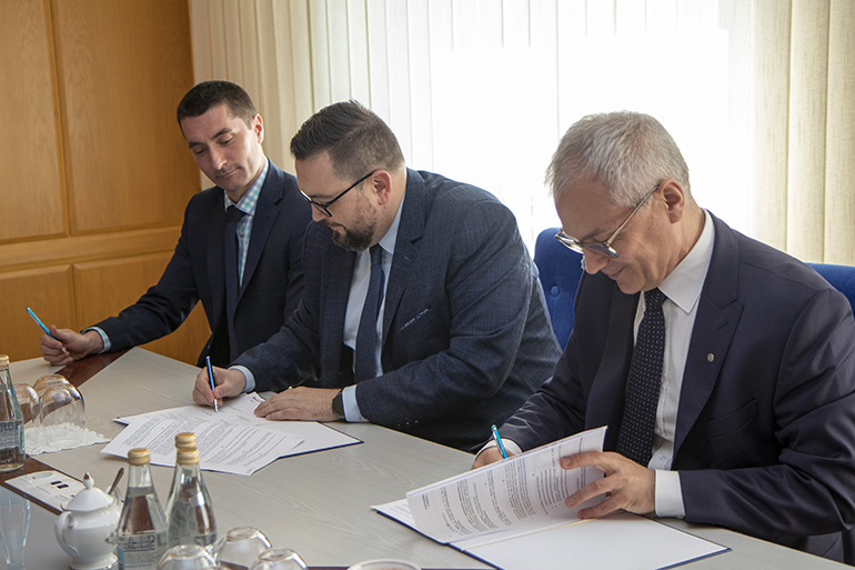 Zdjęcie przedstawia trzech mężczyzn w garniturach siedzących przy stole i podpisujących dokumenty.