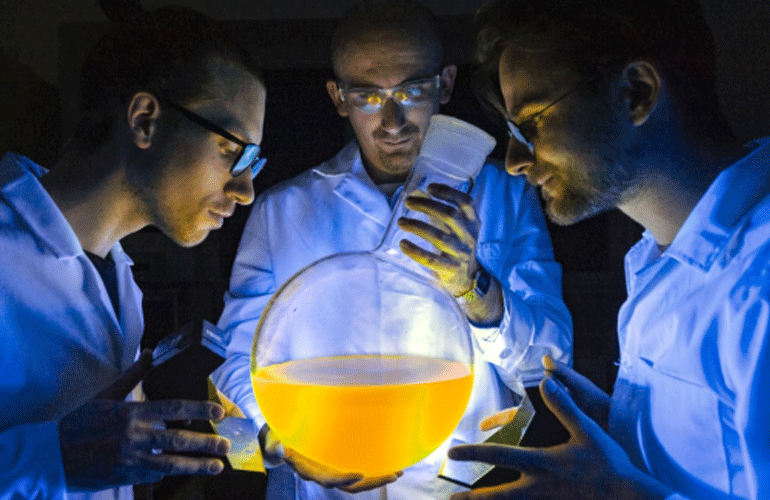 Zdjęcie przedstawia trzech naukowców w fartuchach, pochylających się nad szklanym sprzętem laboratoryjnym..