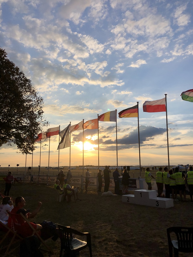 Na zdjęciu widzimy lotnisko na tle zachodzącego słońca. Widać tez fotografów robiących zdjęcia oraz wiszące na masztach flagi różnych państw