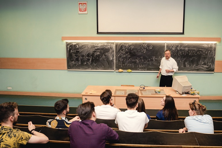 Na zdjęciu widzimy sale wykładową ze studentami, Przed nimi przy tablicy stoi wykładowca