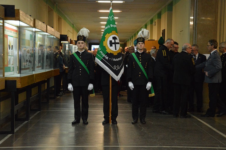 Na zdjęciu widzimy hol wydziału górnictwa. W holu stoją górnicy w galowych mundurach. Na pierwszym planie trzech z nich. Na głowie maja czako. Jeden z nich trzyma uroczysty sztandar.