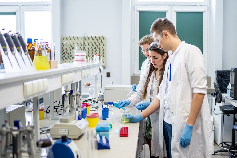 Na zdjęciu widzimy troje w białych fartuchach studentów w laboratorium. Przed nimi stoi blat i półki na których są różne szklane pojemniki