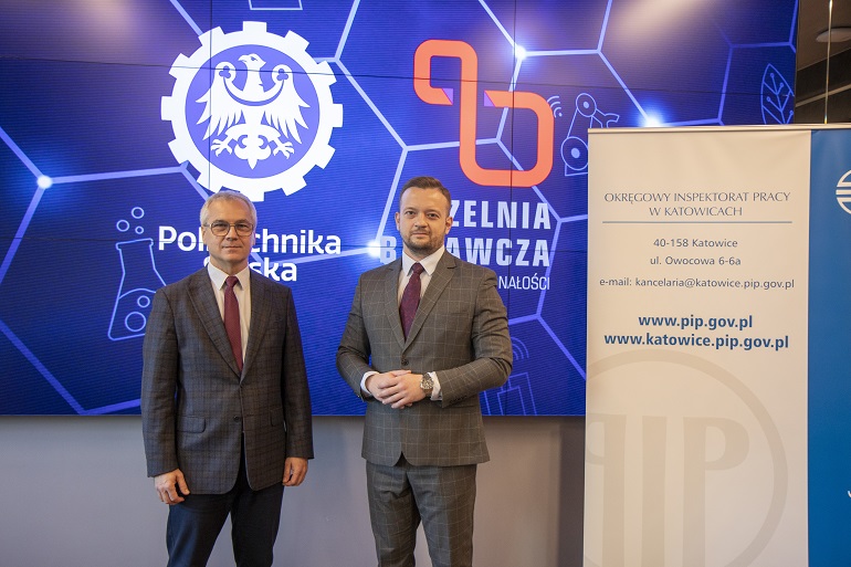 Na zdjęciu widzimy Rektora oraz inspektora pracy stojących na tle dużego ekranu na którym widać logo Politechniki.