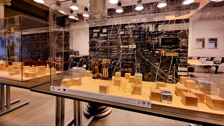 Na zdjęciu widzimy fragment wystawy „Architektoniczny upcycling w teatrze”. Na środku widzimy szklaną gablotę w środku małe tekturowe pudełka