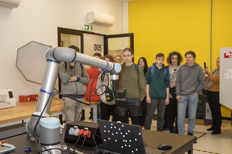 Na zdjęciu widzimy grupę uczniów stojąca w laboratorium. Przed nimi maszyna z ruchomymi ramionami