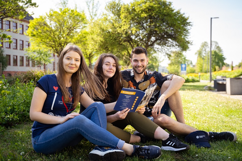 Na zdjęciu widzimy trójkę studentów siedzących na trawie na terenie kampusu