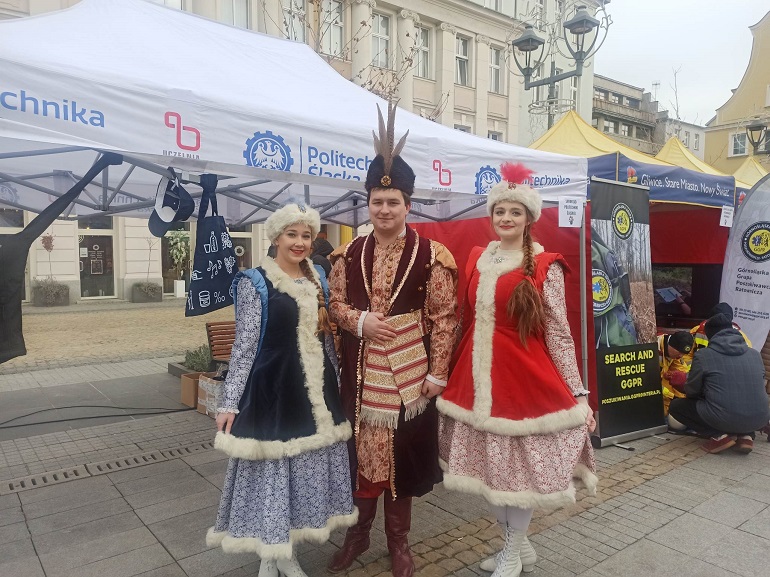 Na zdjęciu widzimy trzech członków Akademickiego Zespółu Tańca Politechniki Śląskiej Dąbrowiacy w strojach szlacheckich. Wszyscy stoją na tle stoiska na rynku