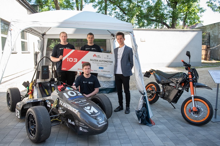 Na zdjęciu widzimy kilku studentów stojących przy bolidzie i motocyklu elektrycznym