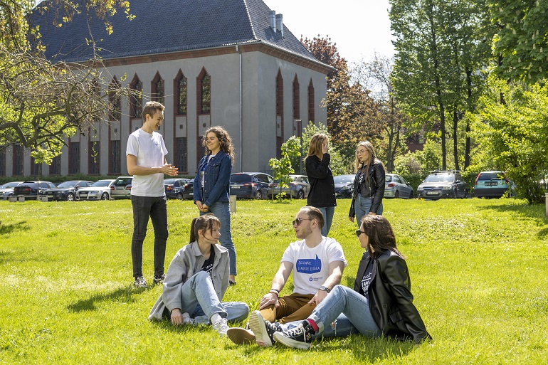 Na zdjęciu widzimy studentów siedzących i stojących na trawie na terenie kampusu uczelni