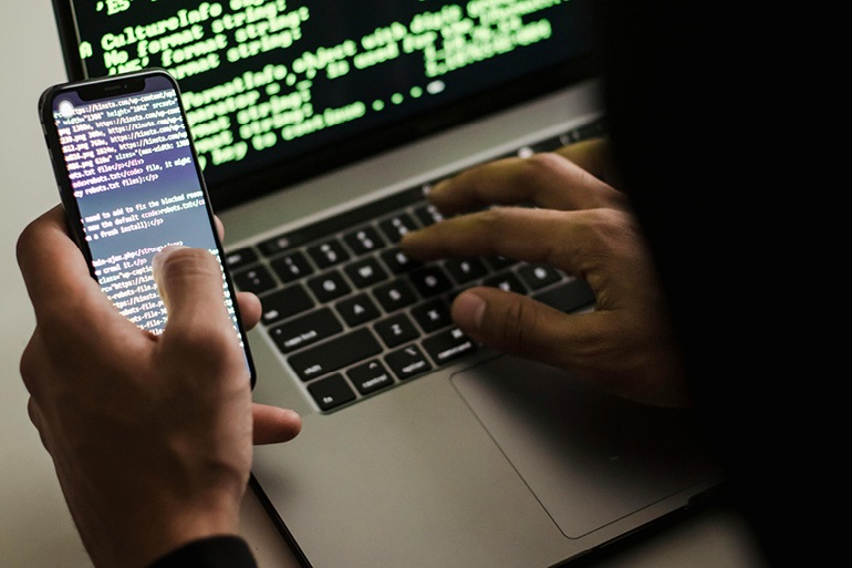 na zdjęciu widzimy otwarty laptop przy którym ktoś siedzi a w drugiej ręce ktoś trzyma smartfon