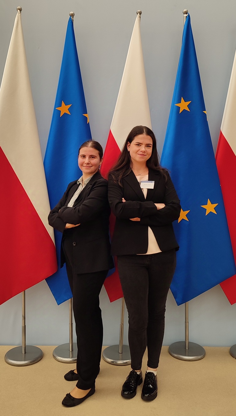 na zdjęciu studentki na tle flag Polski i Unii Europejskiej