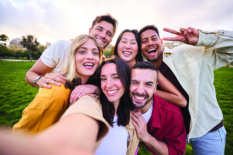 Group of young cheerful multi-ethnic friends taking selfie with mobile phone outdoor. Happy people together smiling and enjoying free time in park. Positive and fun youth relationships in community.