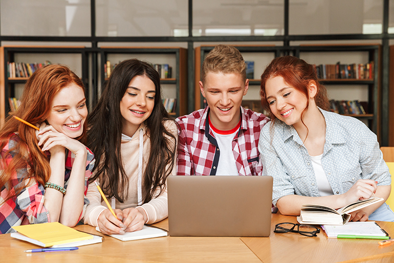 Group of smiling teenagers doing homework while sitting at the library and looking at laptop screen
