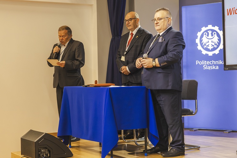 na zdjęciu widzimy trójkę mężczyzn na scenie. Jeden z nich przemawia do mikrofonu